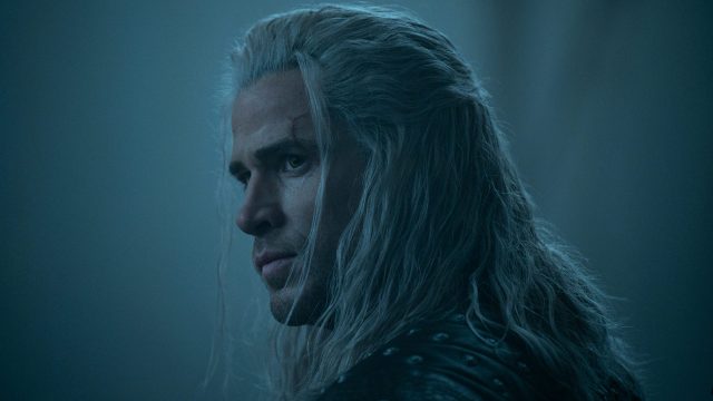 Auf dem Bild zu The Witcher Staffel 4 ist Liam Hemsworth als Geralt von Riva zu sehen. Er trägt langes, silbernes Haar, eine Narbe über dem Auge und eine schwarze Lederrüstung mit Nieten.
