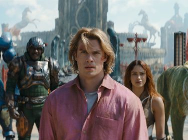 Im Bild zum Masters of the Universe neuer Film ist Adam, gespielt von Nicholas Galitzine, im Vordergrund mit entschlossenem Blick zu sehen. Hinter ihm stehen Teela (Camila Mendes), Man-At-Arms (Idris Elba), Roboto (Kristen Wiig) und der grüne Tiger Cringer. Die Szene wirkt episch und angespannt, mit einer fantasievollen, mittelalterlich-futuristischen Kulisse im Hintergrund.