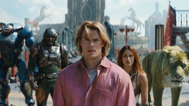 Im Bild zum Masters of the Universe neuer Film ist Adam, gespielt von Nicholas Galitzine, im Vordergrund mit entschlossenem Blick zu sehen. Hinter ihm stehen Teela (Camila Mendes), Man-At-Arms (Idris Elba), Roboto (Kristen Wiig) und der grüne Tiger Cringer. Die Szene wirkt episch und angespannt, mit einer fantasievollen, mittelalterlich-futuristischen Kulisse im Hintergrund.