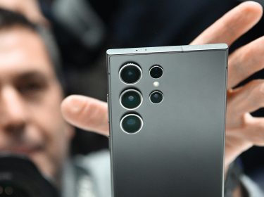 Hand greift nach einem Smartphone im Vordergrund, im Hintergrund das Gesicht eines Mannes