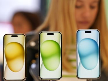 Im Vordergrund mehrere iPhones in der Auslage eines Geschäftes, im Hintergrund guckt eine Frau auf ihr Handy