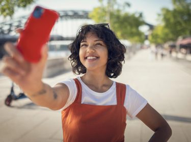 Auf dem Bild in dem Artikel zu den Vodafone CallYa-Tarifen hält eine junge Frau mit lockigen Haaren und orangefarbenem Kleid fröhlich ihr Smartphone in die Höhe, um ein Selfie zu machen. Im Hintergrund ist eine sonnige Fußgängerpromenade mit Bäumen und Cafés zu sehen. Die Szene wirkt lebendig und sommerlich – passend zur frischen Ausstrahlung der Vodafone CallYa-Tarife.