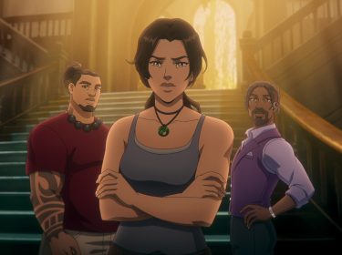 Auf dem Bild aus Tomb Raider: The Legend of Lara Croft stehen Lara Croft, Jonah Maiava und Allen Maldonado vor einer großen, eindrucksvollen Treppe in einem von Licht durchfluteten Gebäude. Lara, in einem grauen Tanktop und mit entschlossenem Blick, steht im Vordergrund mit verschränkten Armen. Hinter ihr sind Jonah, mit Tattoo und rotem Shirt, sowie Allen, in einem lila Anzug mit Ohrstück, zu sehen. Die Szene wirkt angespannt und dynamisch.