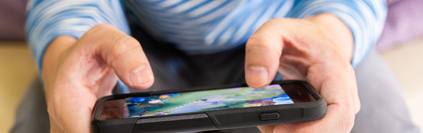 Auf dem Bild zu den besten Handyspielen 2025 hält eine Person mit blau-weiß gestreiftem Shirt ein Smartphone in beiden Händen. Auf dem Bildschirm ist ein farbenfrohes Handyspiel mit grüner Landschaft zu sehen. Die Szene vermittelt eine entspannte Atmosphäre und hebt die Popularität von Mobile Gaming hervor.