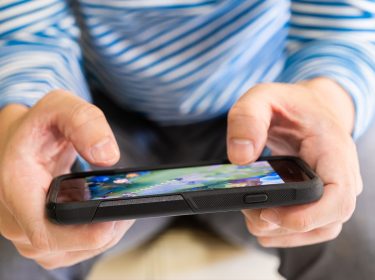 Auf dem Bild zu den besten Handyspielen 2025 hält eine Person mit blau-weiß gestreiftem Shirt ein Smartphone in beiden Händen. Auf dem Bildschirm ist ein farbenfrohes Handyspiel mit grüner Landschaft zu sehen. Die Szene vermittelt eine entspannte Atmosphäre und hebt die Popularität von Mobile Gaming hervor.