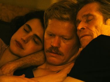 Auf dem Bild zur Kinds of Kindness Filmkritik sieht man drei Personen in inniger Umarmung. Eine junge Frau (Margaret Qualley) schläft, flankiert von zwei nachdenklichen Männern (Jessie Plemons und Willem Dafoe). Die Szene ist in warmen Farbtönen gehalten.