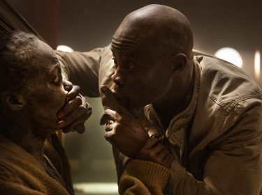 Auf dem Bild zur A Quiet Place: Tag Eins Kritik sieht man Lupita Nyong'o (links), die von Djimon Hounsou (rechts)zum Schweigen gebracht wird, in einer dramatischen und dunklen Szene.
