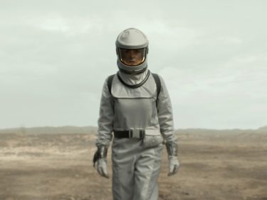 Auf diesem Bild aus Silo Staffel 2ist Rebecca Ferguson in einer beeindruckenden Wüstenlandschaft zu sehen. Sie trägt einen futuristischen Raumanzug in Silbergrau, der sie für die lebensfeindliche Umgebung außerhalb des Silos ausrüstet. Der Anzug ist mit einem dunkelgrauen Gurt und passenden Handschuhen ausgestattet. Ihr Helm verfügt über ein großes, klares Visier, das ihr Gesicht sichtbar macht und ihren ernsten, nachdenklichen Ausdruck hervorhebt. Der Hintergrund zeigt eine trostlose, weite Wüstenlandschaft, die eine karge und unwirtliche Atmosphäre vermittelt, passend zur dystopischen Thematik der Serie.