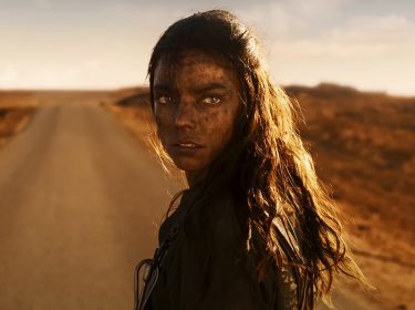 Das Bild im Artikel zu den GigaTV-Highlights im Juli 2024 zeigt Anya Taylor-Joy als Furiosa im Film Furiosa: A Mad Max Saga mit schmutzigem Gesicht und langem, dunklem Haar, die auf einer einsamen, staubigen Straße in einer kargen Wüstenlandschaft steht. Sie schaut ernst und direkt in die Kamera. Im Hintergrund ist der Himmel teils bewölkt.