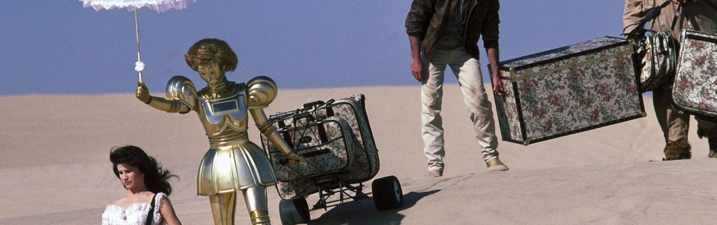 Ein Szenenbild aus Spaceballs zeigt Prinzessin Vespa, ihre Roboterin Dotty, Lone Starr und Waldi auf einer Wanderung durch die Wüste. Die Prinzessin geht voran, um ihre Schulter hängt eine TAsche. Hinter ihr geht die goldene Roboterin und hält einen Sonnenschirm über den Kopf der Prinzessin. Sie zieht einen Gepäckwagen hinter sich hier. Lone Starr folgt ihnen. Er hält mit einer Hand einen sperrigen Koffer. Auf der anderen Seite hilft Waldi beim Tragen des Koffers. Neben den Personen ist nicht als Sand und ein blauer Horizont zu sehen.