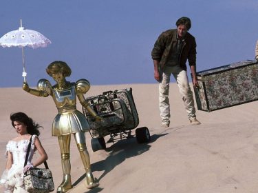 Ein Szenenbild aus Spaceballs zeigt Prinzessin Vespa, ihre Roboterin Dotty, Lone Starr und Waldi auf einer Wanderung durch die Wüste. Die Prinzessin geht voran, um ihre Schulter hängt eine TAsche. Hinter ihr geht die goldene Roboterin und hält einen Sonnenschirm über den Kopf der Prinzessin. Sie zieht einen Gepäckwagen hinter sich hier. Lone Starr folgt ihnen. Er hält mit einer Hand einen sperrigen Koffer. Auf der anderen Seite hilft Waldi beim Tragen des Koffers. Neben den Personen ist nicht als Sand und ein blauer Horizont zu sehen.