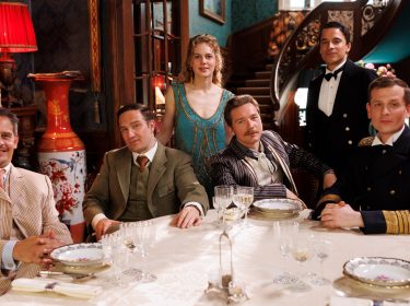 Auf dem Bild aus Dinner For Five sind sechs Personen zu sehen, die in eleganter Kleidung an einem festlich gedeckten Tisch sitzen. Im Hintergrund ist eine aufwendig dekorierte Treppe und ein luxuriös eingerichteter Raum zu erkennen. Die abgebildeten Personen sind Alicia von Rittberg, Kostja Ullmann, Moritz Bleibtreu, Frederick Lau, Jacob Matschenz und Christoph Schechinger.