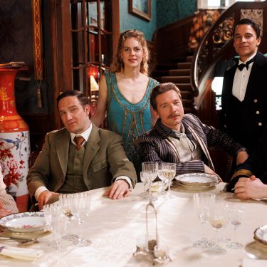 Auf dem Bild aus Dinner For Five sind sechs Personen zu sehen, die in eleganter Kleidung an einem festlich gedeckten Tisch sitzen. Im Hintergrund ist eine aufwendig dekorierte Treppe und ein luxuriös eingerichteter Raum zu erkennen. Die abgebildeten Personen sind Alicia von Rittberg, Kostja Ullmann, Moritz Bleibtreu, Frederick Lau, Jacob Matschenz und Christoph Schechinger.