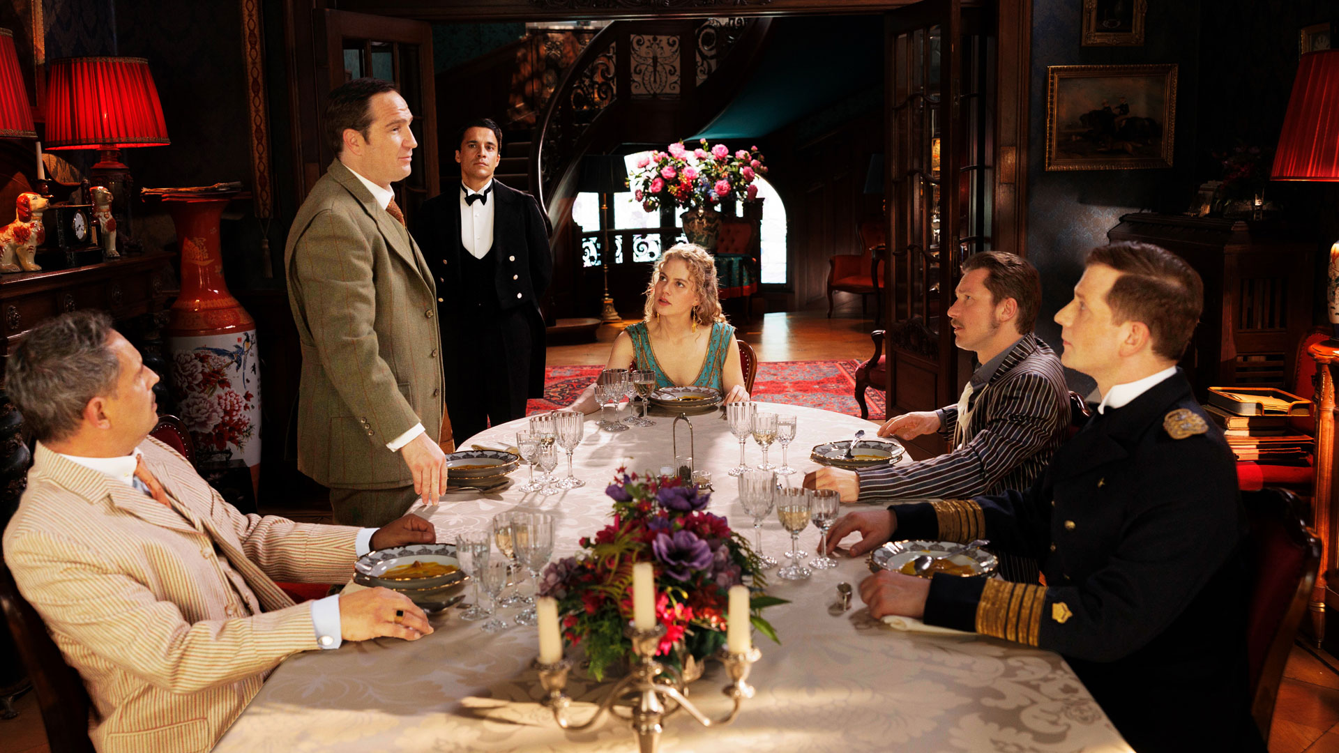 In dieser Szene aus der Prime-Video-Serie Miss Sophie sitzt Miss Sophie (Alicia von Rittberg) mit ihren illustren Gästen – Mr. Pommeroy (Moritz Bleibtreu), Mr. Winterbottom (Frederick Lau), Sir Toby (Jacob Matschenz) und Admiral von Schneider (Christoph Schechinger) – an einer reich gedeckten Tafel. Butler James (Kostja Ullmann) steht im Hintergrund. Die Stimmung ist festlich und nostalgisch.