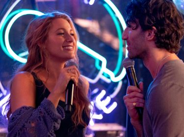 Ein Szenenbild aus dem Nur noch ein einziges Mal-Film zeigt Blake Lively in der linken Bildhälfte als Lily Bloom und Justin Baldony in der rechten Bildhälfte als Ryle. Die beiden schauen sich lächelnd an und haben jeweils ein Mikrofon in der Hand. Im Hintergrund leuchtet ein Karaoke-Symbol in Blau und Lila. Am oberen rechten Bildrand sieht man eine Diskokugel.