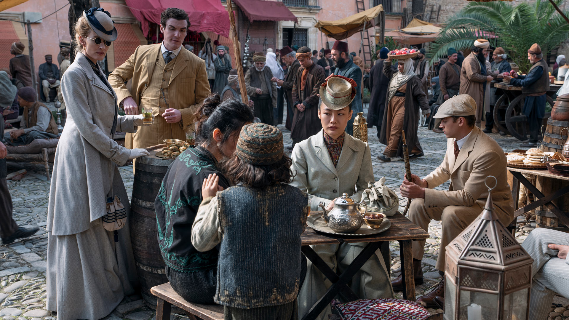 In dieser Szene aus Young Sherlock sind Hero Fiennes Tiffin, Dónal Finn, Zine Tseng und Natascha McElhone in einem historischen Marktplatzsetting zu sehen. Die Figuren tragen detailreiche Kleidung im Stil des späten 19. Jahrhunderts. Zine Tseng sitzt im Zentrum der Szene, umgeben von traditionellen Gegenständen wie einer Teekanne und orientalischen Laternen. Die Atmosphäre ist lebendig und geschäftig.