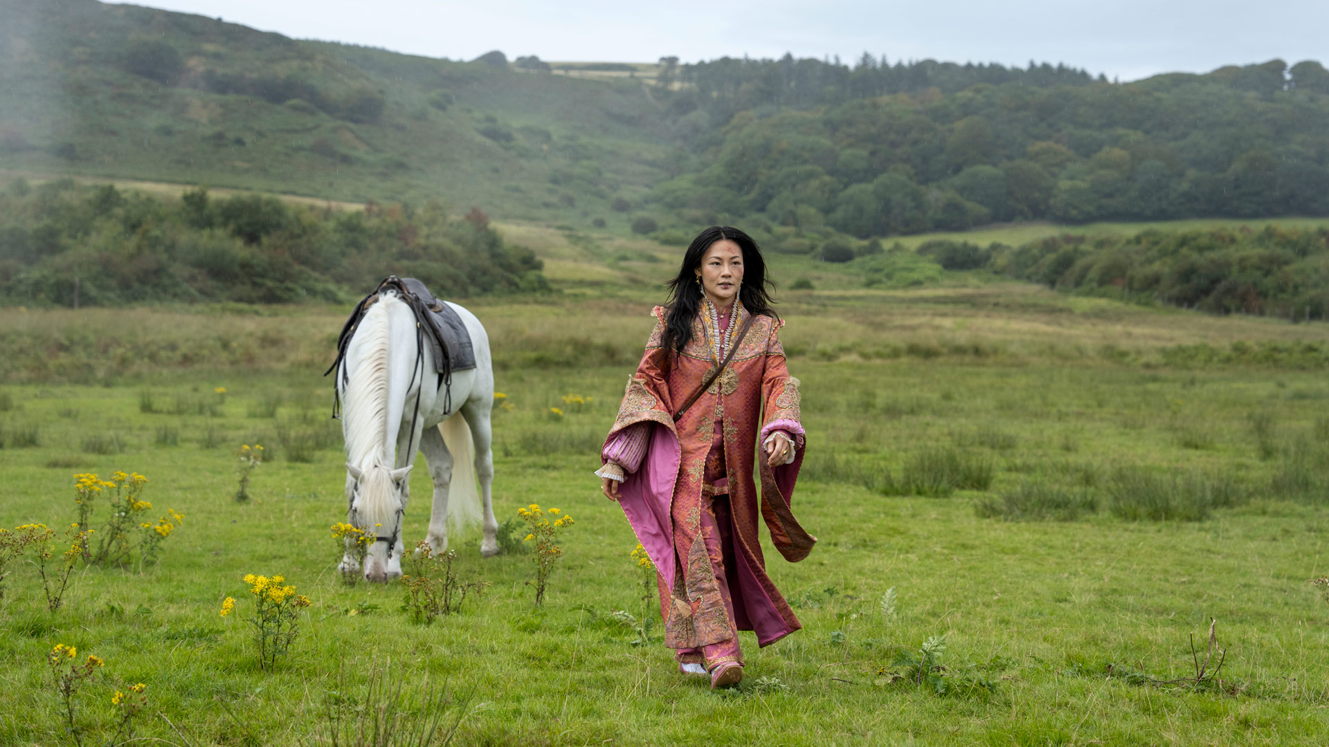 In dieser Szene aus Young Sherlock ist Zine Tseng in einem aufwendig verzierten, rosafarbenen Gewand zu sehen. Sie schreitet über eine weite grüne Wiese, während ein weißes Pferd mit Sattel im Hintergrund grast. Die Landschaft ist ruhig und ländlich, was der Szene eine friedliche, fast mystische Stimmung verleiht.