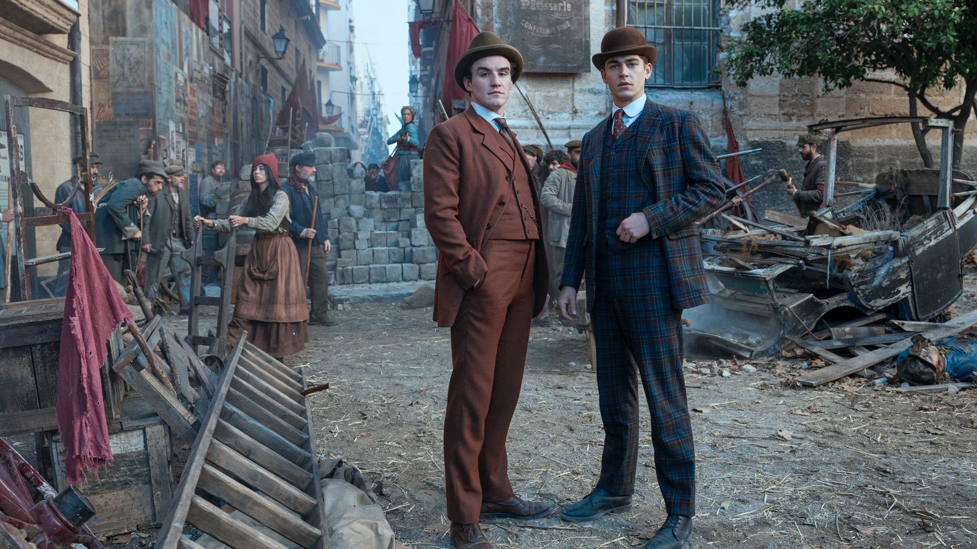 Auf dem Bild aus Young Sherlock stehen Hero Fiennes Tiffin und Dónal Finn im Vordergrund einer aufgewühlten Straßenszene. Beide tragen elegante Anzüge im Stil des frühen 20. Jahrhunderts, Tiffin im karierten Muster, Finn in Braun. Im Hintergrund errichten Bürger eine Barrikade, die Szenerie wirkt angespannt und rebellisch.