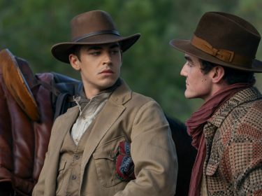 In dieser Szene aus Young Sherlock stehen sich Hero Fiennes Tiffin und Dónal Finn in einer intensiven Gesprächssituation gegenüber. Beide tragen breite Hüte und Outfits im Westernstil, im Hintergrund ist ein Sattel auf einem Pferd zu erkennen. Die Mimik der beiden wirkt ernst und angespannt, was auf eine dramatische Wendung hindeutet.