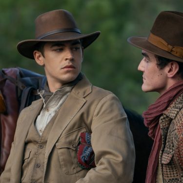 In dieser Szene aus Young Sherlock stehen sich Hero Fiennes Tiffin und Dónal Finn in einer intensiven Gesprächssituation gegenüber. Beide tragen breite Hüte und Outfits im Westernstil, im Hintergrund ist ein Sattel auf einem Pferd zu erkennen. Die Mimik der beiden wirkt ernst und angespannt, was auf eine dramatische Wendung hindeutet.