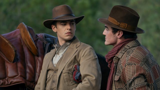 In dieser Szene aus Young Sherlock stehen sich Hero Fiennes Tiffin und Dónal Finn in einer intensiven Gesprächssituation gegenüber. Beide tragen breite Hüte und Outfits im Westernstil, im Hintergrund ist ein Sattel auf einem Pferd zu erkennen. Die Mimik der beiden wirkt ernst und angespannt, was auf eine dramatische Wendung hindeutet.