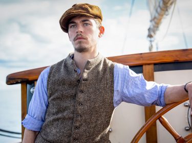 In dem Szenenbild siehst Du den Young Sherlock-Hauptdarsteller Hero Fiennes Tiffin. Er ist auf hoher See und lehnt sich an dem Steuerrad an.