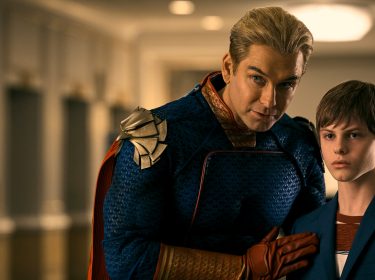 Auf dem Bild zum The Boys Staffel 5 Ausblick sind zwei Charaktere aus einer dramatischen Szene zu sehen. Links steht Homelander, gespielt von Antony Starr, in einem auffälligen Superheldenkostüm mit einer dominanten und beschützenden Haltung. Sein Blick ist intensiv und direkt in die Kamera gerichtet. Rechts von ihm steht Ryan, dargestellt von Cameron Crovetti, ein junger Junge in einem blauen Sakko, der ernst und etwas besorgt aussieht. Die Beziehung zwischen den beiden scheint komplex und intensiv, was durch ihre Körperhaltung und den direkten Augenkontakt unterstrichen wird.