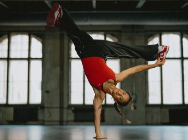 Auf diesem Bild ist die Breakerin Jilou Rasul für die neue Vodafone GO Werbung zu sehen. Sie macht einen Breakdance-Move, indem sie sich auf einer Hand abstützt und ihr Körper in der Luft ist. Mit der linken Hand berührt sie ihren Fuß. Sie trägt eine schwarze Jeans und ein rotes Top.