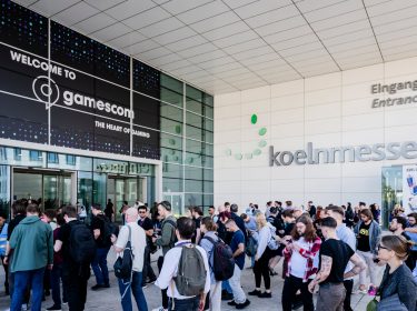Das Bild zeigt den Eingang der Koelnmesse während der gamescom 2024. Vor dem Gebäude versammelt sich eine große Menschenmenge, die auf den Eintritt wartet. Über dem Eingang befindet sich ein großes Schild mit der Aufschrift 