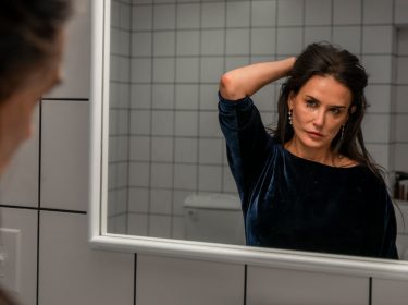Demi Moore steht in The Substance als Elisabeth Sparkle vor dem Badezimmerspiegel. Am linken Bildrand ist verschwommen ihr Gesicht von hinten-rechts zu erkennen sowie ihre Haare. Zentral im Bild sieht man ihr Spiegelbild. Sie scheint geweint zu haben. Ihre Maskara ist unter den Augen verwischt. Sie hält sich die langen, dunklen Haare mit der linken Hand am Hinterkopf aus dem Gesicht und schaut kritisch ihr eigenes Gesicht im Spiegel an. Sie trägt ein samtig-blaues Oberteil und lange Ohrringe. Hinter ihr sind weiße, monotone, quadratische Fliesen der Badezimmerwand zu sehen.