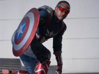 Auf dem Bild im Artikel zu Captain America 4: Brave New World ist Captain America zentral im Bild zu sehen. Vor dem Hintergrund einer neutralen weißen Wand kniet er mit einem Bein auf dem Boden. In der rechten Hand hält er sein Schild, dass so schrägt in die Kamera gerichtet ist. Es ist rund und mit rot-weißen Kreisen verziert. In der Mitte ist ein blauer Kreis mit einem weißen Stern. Auch die UNiform von Captain America passt farblich: sie ist dunkelblau. Auf der Brust des SUperhelden ist ein silberer Stern. Er trägt rote Stiefel und Handschuhe die Teil der metallenen Rüstung sind. Seine Augen werden durch eine festanliegende Brille geschützt mit orangenem durchsichtigem Glas. Er ist schwarz. Captain America blickt konzentriert nach rechts.