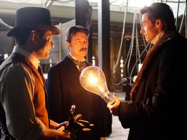 Drei Männer stehen in einer dunklen Werkstatt, einer von ihnen hält eine überdimensionierte Glühbirne in der Hand