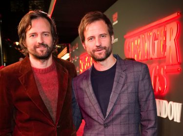 Auf dem Bild zu Something Very Bad Is Going To Happen stehen Matt und Ross Duffer, die Schöpfer von Stranger Things, nebeneinander vor einer Hintergrundwand mit dem Serienlogo zum Theaterstück 