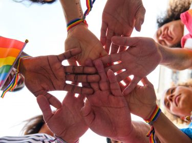 Das Bild zum International Ally Day zeigt eine Gruppe von Menschen, die ihre Hände in der Mitte zusammenschlagen. Sie tragen Armbänder und Kleidung in Regenbogenfarben und halten eine Regenbogenflagge. Die Gesichter der Personen sind fröhlich und motiviert. Das Bild symbolisiert Gemeinschaft und Unterstützung. Keyword: International Ally Day.