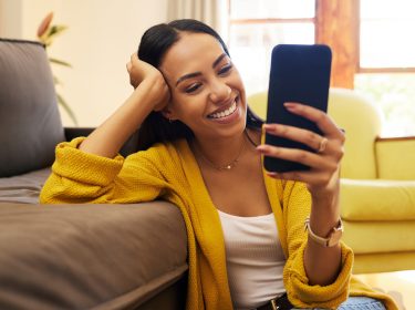 Auf dem Bild zur Butterflies App sitzt eine Frau auf dem Boden vor einem Sofa und schaut lächelnd auf das Smartphone in ihrer Hand.