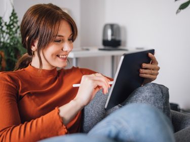 Auf dem Bild im Artikel dazu, wie Du Vodafone digital erreichen kannst, sitzt eine junge Frau entspannt auf einem Sofa und nutzt ein Tablet mit einem Eingabestift.