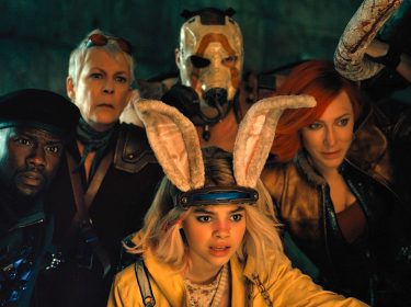 Im Bild zur Borderlands-Filmkritik sind 5 Hauptcharaktere zu sehen. Sie alle schauen erstaunt und neugierig in dieselbe RIchtung. Der Hintergrund ist dunkel, die Gesichter der Figuren werden von einer warmen Lichtquelle vor ihnen angestrahlt. Links im Bild befindet sich Roland (Kevin Hart). Erträgt eine dunkle Uniform mit schwarzer Soldaten-Kappe. Zentral im Bild und vor den anderen hockt Tiny Tina (Ariana Greenblatt). Sie hat eine gelbe Jacke an. Ihre Haare sind schulterlang und blond gefärbt. Auf ihrem Kopf trägt sie eine Art Haarreif mit plüschigen Hasenohren. Rechts im Bild ist Lilith (Cate Blanchett) zu sehen. Sie trägt dunkle Klamoten. Ihre Haare sind rot gefärbt. In der Mitte hinten steht Krieg (Florian Monteanue). Sein Körper ist von Tiny Tina verdeckt. Sein Gesicht ist ebenfalls nicht zu erkennen, denn er trägt eine weiße Maske mit einem orangenen Bogen darauf. Im Bild links von ihm ist Tannis (Jamie Lee Curtis) Gesicht zu sehen. Sie hat kurze graue Haare und trägt eine Art Sonnenbrille auf dem Kopf.