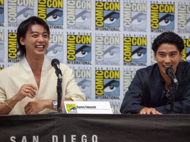 Auf diesem Bild von der Comic-Con sind die Schauspieler Ryoma Takeuchi (links) und Kento Kaku (rechts) bei einem Panel zu sehen. Beide lachen und wirken entspannt, während sie vor einem Hintergrund mit Comic-Con-Logos sitzen. Takeuchi trägt ein weißes Hemd, während Kaku in einem dunklen Anzug gekleidet ist. Sie sprechen über ihre Rollen in der 