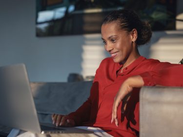 Auf dem Bild im Artikel zu Vodafone connect Festnetztest 2024 siehst Du eine Frau, die lächelnd in ihrem Wohnzimmer an ihrem Laptop sitzt. Sie trägt ein rotes Oberteil.