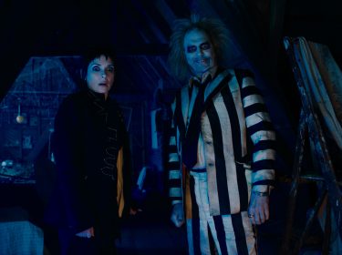Auf diesem Bild zur Beetlejuice Beetlejuice Kritik stehen Winona Ryder als Lydia und Michael Keaton als Beetlejuice nebeneinander in einem düsteren, von Spinnweben bedeckten Dachboden. Lydia trägt schwarze Kleidung und blickt überrascht in die Kamera, während Beetlejuice in seinem markanten schwarz-weiß gestreiften Anzug grinst. Die Szene ist in kaltes, blaues Licht getaucht, was eine geheimnisvolle und leicht unheimliche Atmosphäre erzeugt. Im Hintergrund sind ein Leitergerüst und Abdeckungen zu erkennen.