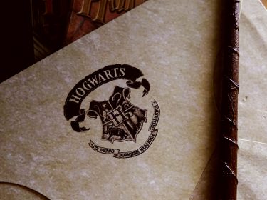 Auf dem Bild ist ein Briefumschlag zu sehen, der das Wappen der Hogwarts-Schule für Hexerei und Zauberei trägt. Neben dem Umschlag liegt ein Zauberstab, der leicht schräg auf dem Papier ruht. Das Wappen zeigt die vier Häuser der Schule und den Schriftzug 