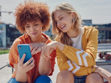 Auf dem Bild in dem Artikel zu Vodafone Young Tarifen sitzen zwei junge Frauen im Freien auf einer Fläche. Sie schauen gemeinsam auf ein Smartphone mit blauer Hülle.