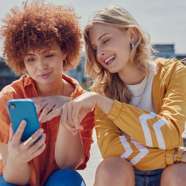 Auf dem Bild in dem Artikel zu Vodafone Young Tarifen sitzen zwei junge Frauen im Freien auf einer Fläche. Sie schauen gemeinsam auf ein Smartphone mit blauer Hülle.