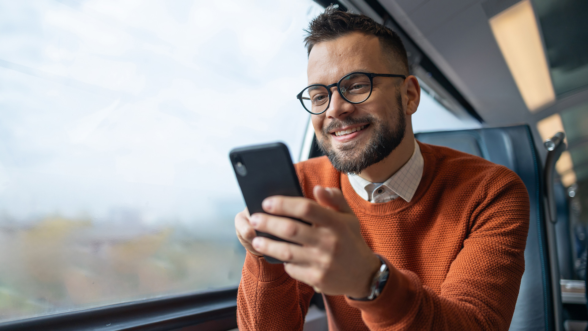 Auf dem Bild in dem Artikel zu den GigaMobil-Tarifen sitzt ein junger Mann mit Brille und Bart in einem Zug am Fenster. Er trägt einen orangefarbenen Pullover und lächelt, während er auf sein Smartphone schaut.