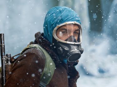 Auf dem Bild vom Eternauta-Netflix-Artikel ist die Figur Juan aus der Netflix-Show Eternauta zu sehen. Er trägt eine dicke Winterjacke mit Fellkragen, eine blaue Kapuze und eine Gasmaske, die ihn vor den gefährlichen Bedingungen in der apokalyptischen Welt schützt. Auf seinem Rücken trägt er einen Rucksack und eine Waffe, bereit, sich den Gefahren seiner Umgebung zu stellen. Schneeflocken fallen um ihn herum und die eisige Landschaft im Hintergrund verstärkt die düstere, bedrückende Atmosphäre, die die verzweifelte Situation in Eternauta widerspiegelt.
