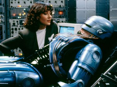 In diesem Szenenbild aus *RoboCop 2* sehen wir die Figur RoboCop in einem High-Tech-Labor auf einer Liege, während er von einer Frau in einem weißen Oberteil und schwarzen Blazer betreut wird. Die Frau schaut aufmerksam auf RoboCop, der in seiner ikonischen metallisch-blauen Rüstung daliegt. Kabel und Schaltkreise sind an seinen Körper angeschlossen, während im Hintergrund eine Vielzahl an Monitoren und Schaltflächen aufleuchtet, was die futuristische und düstere Atmosphäre unterstreicht. Diese Szene zeigt RoboCop in einer ruhenden Position, vermutlich während einer Wartungs- oder Reparatursitzung, und verstärkt die technologische und menschliche Dualität der Figur – ein Thema, das auch in einem modernen RoboCop-Reboot spannend wäre.