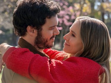 Auf dem Bild aus der Serie „Nobody Wants This“ sind die Charaktere Joanne und Noah in einer liebevollen Umarmung zu sehen. Sie schauen sich mit zärtlichen Blicken an, während Joanne ihre Arme um Noah legt. Beide lächeln sich sanft an, was eine tiefe Verbundenheit zwischen ihnen vermittelt. Joanne trägt einen roten Pullover, und im Hintergrund blühen Bäume in einer friedlichen, sonnigen Parklandschaft, was die warme und romantische Atmosphäre der Szene unterstreicht.