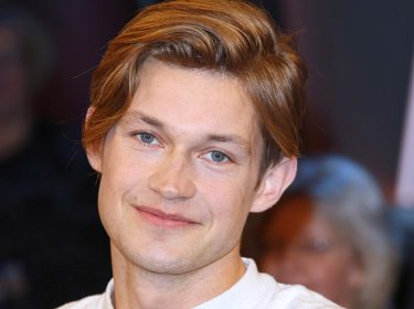 Auf dem Bild ist der deutsche Schauspieler Damian Hardung zu sehen, wie er in die Kamera lächelt. Er hat hellbraune Haare, die leicht zur Seite gestylt sind, und blaue Augen. Sein Gesichtsausdruck wirkt freundlich und entspannt. Der Hintergrund ist unscharf, sodass der Fokus auf seinem Gesicht liegt.