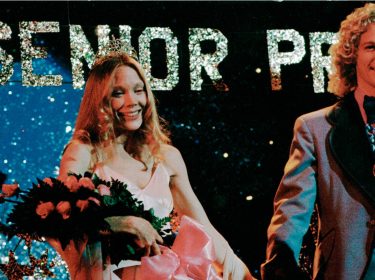 Auf dem Bild zur Serie Carrie sieht man die gekrönte Carrie, gespielt von Sissy Spacek, auf ihrem Abschlussball. Sie trägt eine Tiara und hält einen Blumenstrauß mit rosafarbenen Rosen, während sie lächelnd neben ihrem Ballkönig steht. Im Hintergrund leuchtet ein großes 