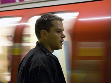 Auf dem Bild ist Jason Bourne, gespielt von Matt Damon, in einer U-Bahn-Station zu sehen. Bourne steht seitlich im Profil, fokussiert und konzentriert, während hinter ihm ein vorbeirasender roter U-Bahn-Wagen Bewegungsunschärfe erzeugt. Die Szene vermittelt Spannung und Dynamik, passend zu Bournes actionreichem Charakter und seiner ständigen Flucht.